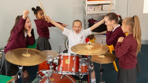 A young female pupil sat at a drum kit with her arms raised and holding a rock star pose, while her classmates make silly faces at her, stood around her in a semi circle 