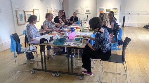 An room with art on the walls and a table and chairs in the middle where a group of women are sitting and taking part in a monoprinting workshop. Lots of art materials are scattered across the table