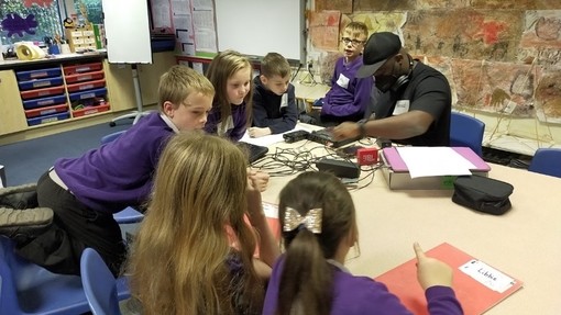 Children wearing purple jumpers and an adult wearing a black t-shirt and black cap sit round a table with audio recording equipment on it